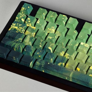 Könnte beinhalten: Eine mechanische Tastatur mit individuellen Tastenkappen in einem Farbverlauf von dunklem Waldgrün zu hellem Lindgrün. Die Tastenkappen sind mit gelben Blumenmustern und abstrakten Pinselstrich-Texturen auf der Oberfläche verziert.