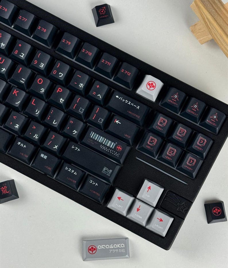 May include: A black mechanical keyboard with dark keycaps featuring red Japanese characters and symbols. The spacebar displays a barcode and a red circular logo. A separate light gray keycap with the Arasaka logo and text is placed below.
