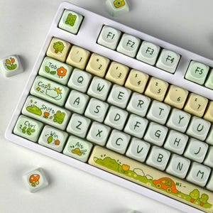 Könnte beinhalten: Eine weiße mechanische Tastatur mit individuellen Tastenkappen in Hellgrün, Gelb und Weiß. Die Tasten zeigen Buchstaben, Zahlen und verschiedene naturbezogene Illustrationen wie Frösche, Blumen, Wolken und ein Auto. Die Leertaste zeigt eine Landschaft mit einem Auto und Bäumen, und der Text "Hi-" ist auf der rechten Seite aufgedruckt.