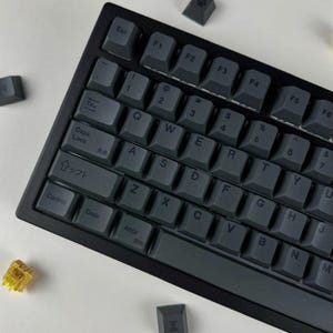 Könnte beinhalten: Eine schwarze mechanische Tastatur mit dunkelgrauen Tastenkappen und weißer Beschriftung. Das Layout umfasst Standard-QWERTY-Tasten sowie spezifische japanische Schriftzeichen auf den Shift-, Code- und Caps-Lock-Tasten. Einzelne Tastenkappen liegen daneben.