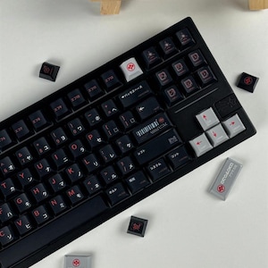 May include: A black mechanical keyboard featuring custom keycaps with red Japanese characters and English letters. The layout includes grey arrow keys and a grey escape key with a red logo. Several loose keycaps are scattered around the keyboard.