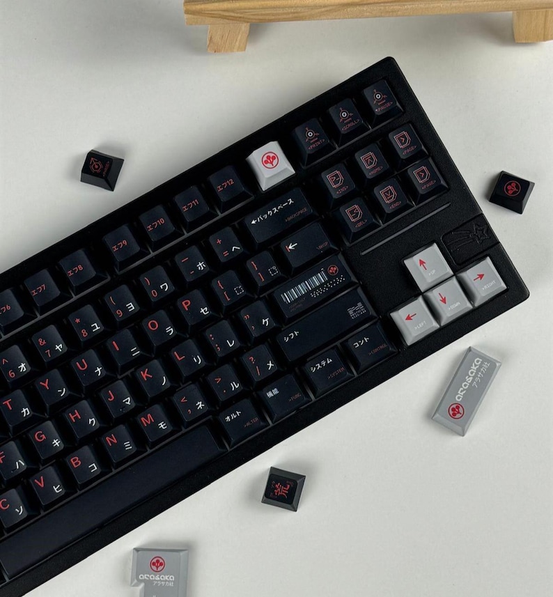 May include: A black mechanical keyboard featuring custom keycaps with red Japanese characters and English letters. The layout includes grey arrow keys and a grey escape key with a red logo. Several loose keycaps are scattered around the keyboard.