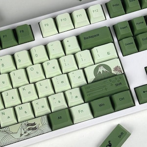 May include: A mechanical keyboard featuring a colour scheme of light and dark green keycaps. The right shift key displays a graphic of a mountain and a tree, while the spacebar features a wave pattern. Several keys include Japanese hiragana characters.