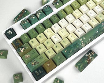 May include: A mechanical keyboard with a white frame and a custom keycap set featuring a gradient of green and cream colors. The keycaps are decorated with botanical illustrations, including lily pads, flowers, and birds, with standard QWERTY lettering.