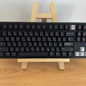 May include: A black mechanical keyboard featuring red alphanumeric characters and Japanese katakana sub-legends. The layout includes a white escape key, white arrow keys, and a barcode graphic with the text YAMAZAK on the right side of the board.