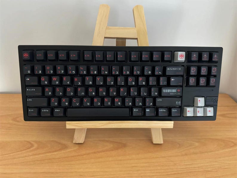 May include: A black mechanical keyboard featuring red alphanumeric characters and Japanese katakana sub-legends. The layout includes a white escape key, white arrow keys, and a barcode graphic with the text YAMAZAK on the right side of the board.