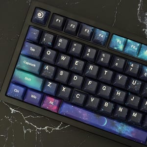 Könnte beinhalten: Eine mechanische Tastatur mit individuellen Tastenkappen in einem dunklen Weltraum-Design. Die Tasten zeigen weiße Buchstaben, Zahlen und Symbole, wobei einige Tasten Nebel-Muster, Sterne und ein Mondsichel-Symbol aufweisen. Die Leertaste zeigt einen Planeten.