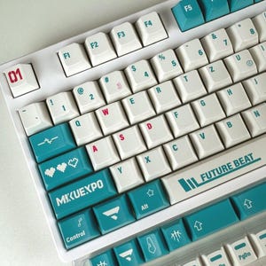 Può includere: Una tastiera meccanica bianca con tasti color verde acqua e bianchi. I tasti presentano scritte color verde acqua, con tasti specifici che mostrano il testo MIKUEXPO, FUTURE BEAT e 01. Alcuni tasti includono icone di cuori pixelati e note musicali.