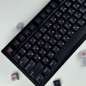 May include: A black mechanical keyboard featuring dark keycaps with red lettering. The keys display both Latin characters and Japanese Katakana symbols. Several loose, light gray and black decorative keycaps with red icons are scattered nearby.