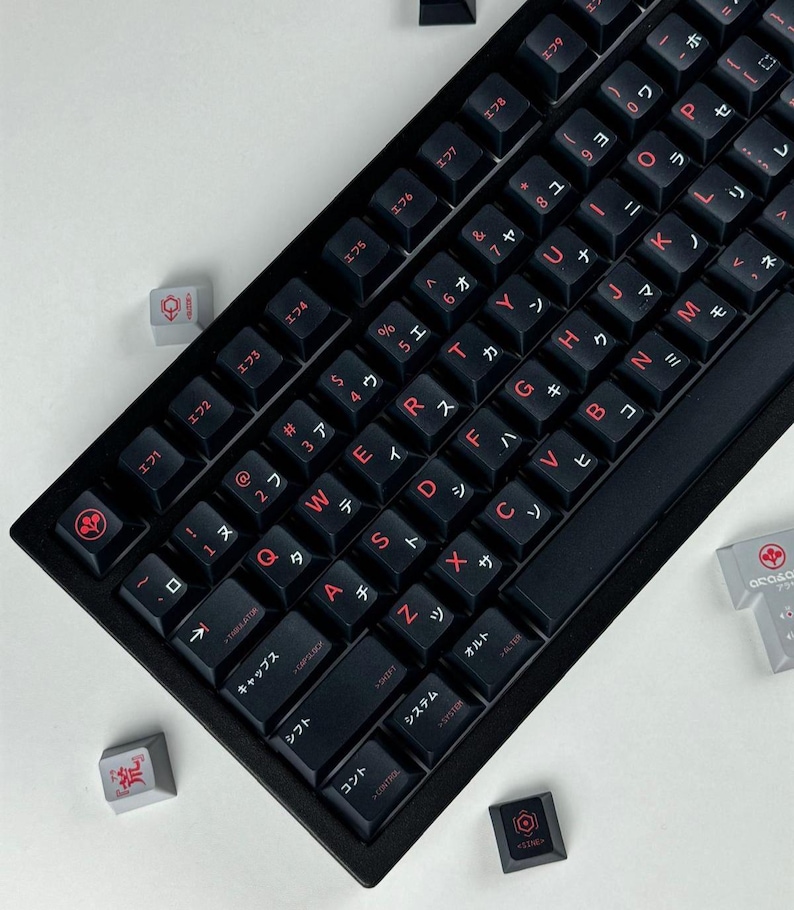 May include: A black mechanical keyboard featuring dark keycaps with red lettering. The keys display both Latin characters and Japanese Katakana symbols. Several loose, light gray and black decorative keycaps with red icons are scattered nearby.