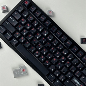May include: A black mechanical keyboard featuring red alphanumeric characters and Japanese katakana legends on each keycap. Several loose gray and black keycaps with red symbols and text are scattered around the main keyboard unit.