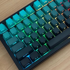 Könnte beinhalten: Eine mechanische Tastatur mit schwarzem Rahmen und einem Farbverlauf auf den Tastenkappen, der von Türkis oben links bis Dunkelgrau unten rechts reicht. Die Tasten sind mit einer lebendigen RGB-Beleuchtung hinterleuchtet, die die Buchstaben hervorhebt.
