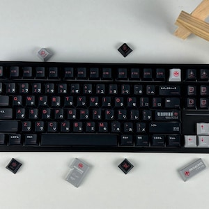 May include: A black mechanical keyboard featuring dark keycaps with red English letters and Japanese katakana characters. The layout includes a white backspace key, light gray arrow keys, and several loose keycaps scattered around the frame.
