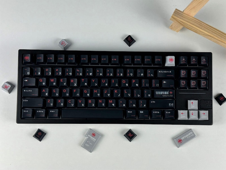 May include: A black mechanical keyboard featuring dark keycaps with red English letters and Japanese katakana characters. The layout includes a white backspace key, light gray arrow keys, and several loose keycaps scattered around the frame.