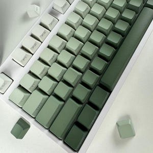 Könnte beinhalten: Eine mechanische Tastatur mit einem Farbverlauf, der von hellem Mintgrün zu tiefem Waldgrün über die Tastenkappen reicht. Die Tastatur hat einen weißen Rahmen, und zwei lose Tastenkappen sind außerhalb des Haupttastaturbereichs positioniert.