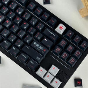 May include: A black mechanical keyboard featuring dark keycaps with red Japanese characters and English sub-legends. The layout includes a white backspace key, light gray arrow keys, and a rectangular gray spacebar with the Arasaka logo and text.