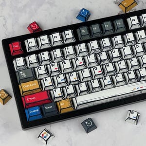 May include: A mechanical keyboard featuring white keycaps with black comic-style outlines and hatching. The layout includes a red Escape key, a yellow Caps Lock key, a red Shift key, and blue Control and Super keys. Extra loose keycaps are scattered.