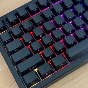 Könnte beinhalten: Eine schwarze mechanische Computertastatur mit mattschwarzen Tastenkappen und weißer Beschriftung. Die Tastatur wird durch eine lebendige RGB-Hintergrundbeleuchtung erhellt, die in Rot-, Lila- und Orangetönen unter den Tasten leuchtet.