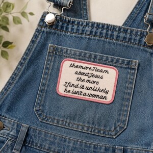 May include: Blue denim overalls featuring a rectangular patch on the chest pocket. The patch has a cream-colored background with a pink border and black embroidered text that reads: the more I learn about Jesus the more I find it's unlikely he isn't a woman.