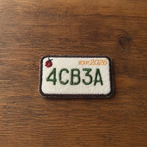 May include: A rectangular embroidered patch with a white background and a dark brown border. The patch features the green text 4CB3A, a small red ladybug icon in the top left corner, and the orange text EXP 2026 in the top right corner.