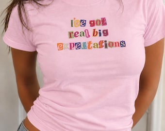 May include: A light pink short-sleeved t-shirt featuring the text "i've got real big expectations" printed in a multi-colored ransom note style with individual letters in various fonts and colors against a solid pastel pink background.