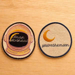May include: Two circular embroidered patches. The left patch features an astronaut helmet with a yellow crescent moon and the text "I'm an astronaut" in black. The right patch shows a yellow crescent moon and the text "you're the moon" in black.