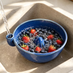 Könnte beinhalten: Ein tiefblaues Beeren-Sieb aus Keramik mit einem zentralen Kegel und einem seitlichen Ausguss für den Wasserabfluss. Die Schale ist mit frischen Blaubeeren und roten Erdbeeren gefüllt, die unter fließendem Wasser abgespült werden.