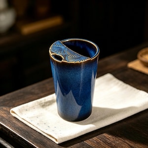 May include: A deep blue ceramic tumbler with a glossy finish and a semi-circular rim feature. The top edge displays a textured, fan-like embossed pattern in a metallic bronze tone, contrasting with the smooth, dark blue glazed body of the vessel.
