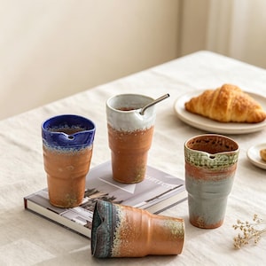 May include: Four ceramic tumblers with a rustic, unglazed terracotta base and a glazed upper rim in blue, white, green, and dark teal. Each tumbler features a small pouring spout at the rim. One tumbler contains a thin, silver-coloured metal straw.