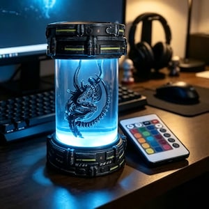 May include: A cylindrical lamp featuring a black base and top with yellow accents, containing a clear chamber illuminated by a bright blue light. Inside the chamber is a dark, intricate, organic-shaped sculpture. A multi-colored remote control is nearby.