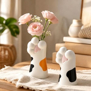 May include: Two white ceramic vases shaped like cat paws with black and orange spots and pink toe pads. The taller vase holds three light pink flowers and small white filler flowers. The design features rounded toes and a smooth, glossy finish.
