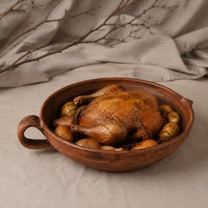 Può includere: Una teglia rotonda in argilla marrone rustica con un manico laterale, contenente un pollo intero arrosto dorato circondato da diverse piccole patate cotte. Il recipiente presenta una finitura strutturata e terrosa con un bacino profondo.