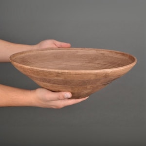 May include: A large, wide, shallow bowl crafted from light brown clay with a visible wood grain texture etched into the surface. The bowl features a smooth, tapered rim and a consistent earthy tone throughout its circular, open design.