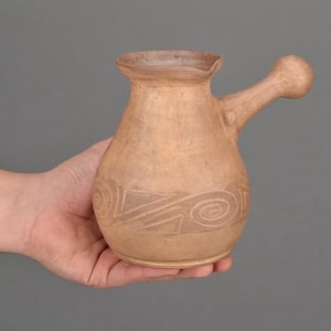 May include: A light brown clay coffee pot with a rounded body, a wide flared rim, and a single side handle ending in a knob. The lower portion of the pot features a carved band of repeating spiral and geometric patterns in a matte finish.