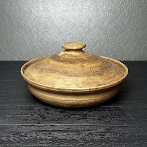 May include: A round, shallow ceramic cooking pot with a matching lid, featuring a light brown, mottled finish and subtle, hand-etched spiral and linear patterns across the exterior surface of both the base and the lid, including a knob handle.