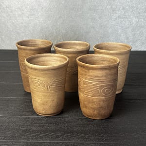 May include: A set of five cylindrical brown clay cups with a matte finish. Each cup features a hand-carved spiral and geometric pattern encircling the middle section. The vessels have a tapered shape with a smooth rim and a consistent earthy tone.