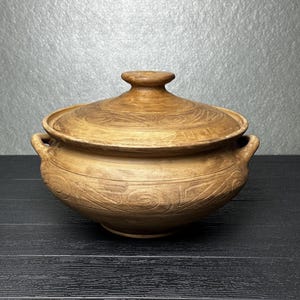 Può includere: Una pentola in argilla di colore marrone chiaro di forma rotonda con coperchio abbinato. Il recipiente presenta due piccole maniglie laterali ed è decorato con motivi circolari a spirale incisi sul corpo e sul coperchio. La superficie ha una texture opaca.