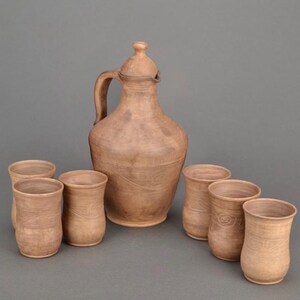 May include: A set of six terracotta-colored clay vessels consisting of one large pitcher with a lid and handle, accompanied by five matching drinking cups. Each piece features a smooth, matte finish with subtle, swirling, organic texture patterns.