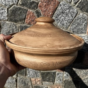 May include: A round, light brown clay cooking pot with a matching lid. The exterior surface of both the pot and the lid features a series of carved, swirling spiral patterns that wrap around the circumference of the vessel in a repeating design.