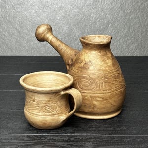 Könnte beinhalten: Eine beige Keramik-Kaffeekanne mit langem Griff und eine passende Keramiktasse. Beide Teile haben eine braun marmorierte Oberfläche und geprägte Spiralmuster. Die Kanne hat einen breiten Boden und einen schmalen Hals, die Tasse einen Henkel.
