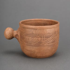 May include: A terracotta-colored clay mug with a rounded, ball-shaped handle. The exterior surface features a hand-carved decorative band consisting of swirling spiral patterns and geometric lines encircling the middle of the vessel.