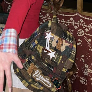 May include: A plaid backpack featuring a mix of yellow, blue, and black fabric patterns. The bag is decorated with two white star-shaped pins, small plush animal figures, and a patch with the white cursive text Vernichter stitched on the front.