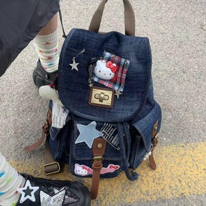 May include: A dark blue denim backpack featuring a flap closure with a gold-tone metal buckle. The bag is decorated with a Hello Kitty character pin, a red and blue plaid fabric patch, a light blue star patch, and a Hello Kitty sticker.