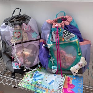 May include: Two drawstring backpacks featuring heart-shaped mesh front pockets. The left bag is gray and lavender with a star patch. The right bag is teal, purple, and pink with a 'GOOD LUCK' patch. Both bags are decorated with various charms.