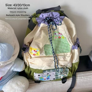 May include: A nylon drawstring backpack measuring 43 by 30 by 13 cm. The bag features olive green and cream panels, a purple drawstring top, and a Hello Kitty graphic patch. It includes a star-shaped applique, a green checkered patch, and a metal charm.