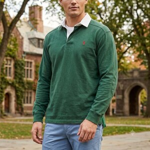 May include: A long-sleeve forest green rugby shirt featuring a white collar, a button placket, and a small red embroidered logo on the left chest. The shirt is paired with light blue pants, creating a classic look with ribbed cuffs.