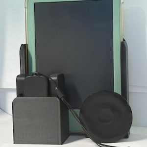 May include: A black 3D-printed plastic stand holding a tablet with a light green case. The stand features a vertical slot for the tablet, a smaller compartment for a black rectangular device, and a circular black disc attached by a black lanyard.