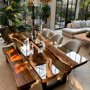 Könnte beinhalten: Ein Esstisch aus Massivholz mit Naturkante, einer hochglänzenden Harzbeschichtung und einer passenden Holzbank. Der Tisch zeigt eine natürliche Holzmaserung in Dunkelbraun, steht auf schwarzen Metallbeinen und ist von hellen Polsterstühlen umgeben.