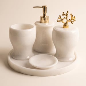 Op de afbeelding: Een badkameraccessoireset van wit marmer bestaande uit een zeepdispenser met een gouden pomp, een beker, een potje met deksel en een gouden koraalvormige knop, en een klein zeepbakje, alles geplaatst op een bijpassend rond marmeren dienblad.