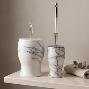 Peut inclure: Un pot en marbre blanc avec un couvercle surmonté d'une poignée en forme de plume argentée et un porte-brosse de toilette en marbre blanc assorti avec un manche en chrome poli. Les deux objets présentent des veines gris foncé sur la pierre.